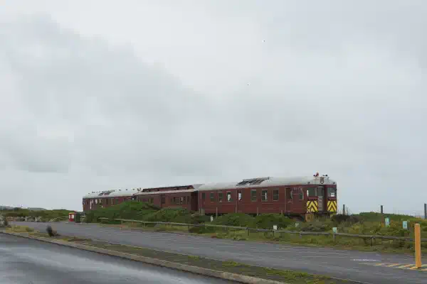 A diesel locomotive heading to the right of the scene, coastal dunes with greenery in the foreground.