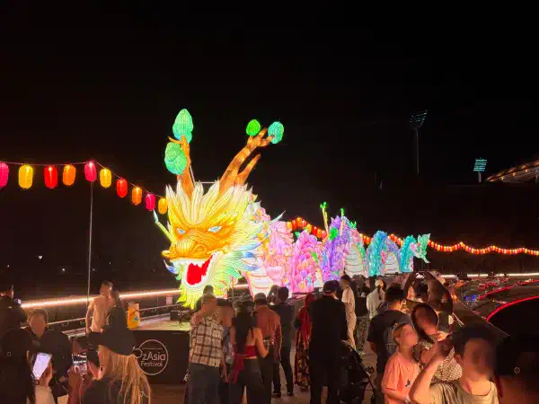A Chinese dragon installation atop a bridge at night-time. It is lit from within. It has many vibrant colours. Many people are walking near it