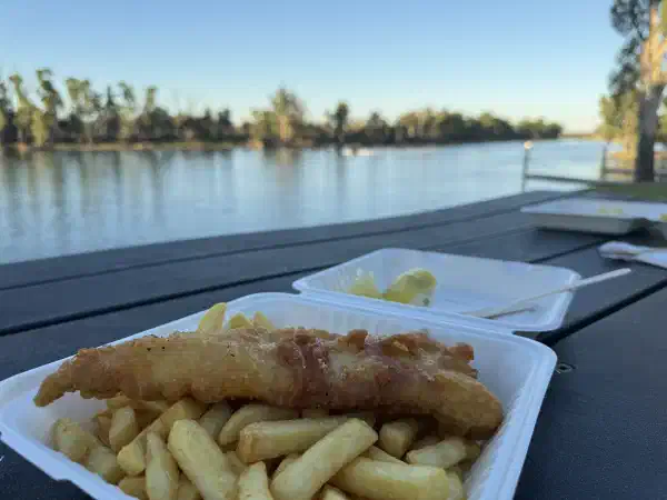 fish and chips in a styrofoam container, on a table. The river is in the background