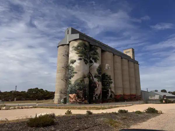 a very large grain silo, with a colourful mural painted on the outside, trees with dogs and sheep at the base