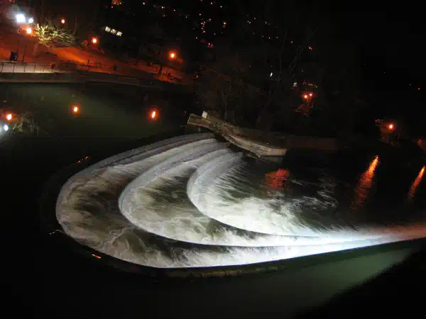 A river, with tiers of human-made waterfalls in tiers, in a tight curve shape. It is nighttime but lit up