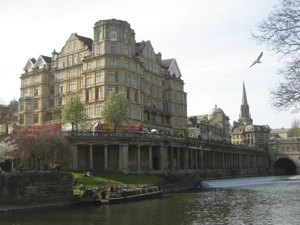 View of a river from ground level, in the background is an embankment with many large buildings. On the river is a longboat