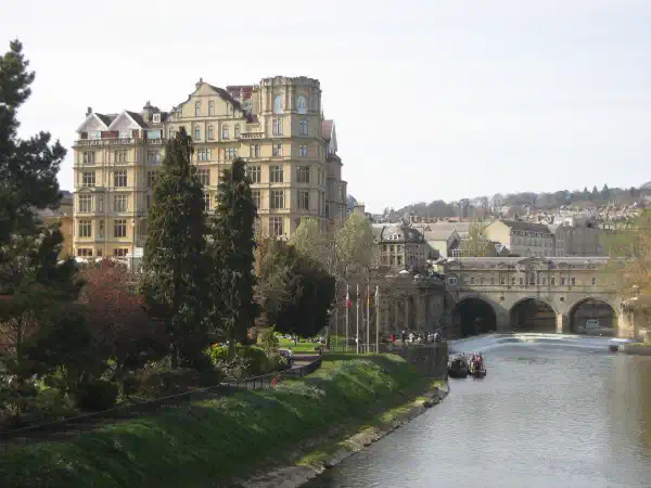 Riverside view, a grassy park is on the far bank with old buildings in the background. An arched bridge crosses the bridge in the distance