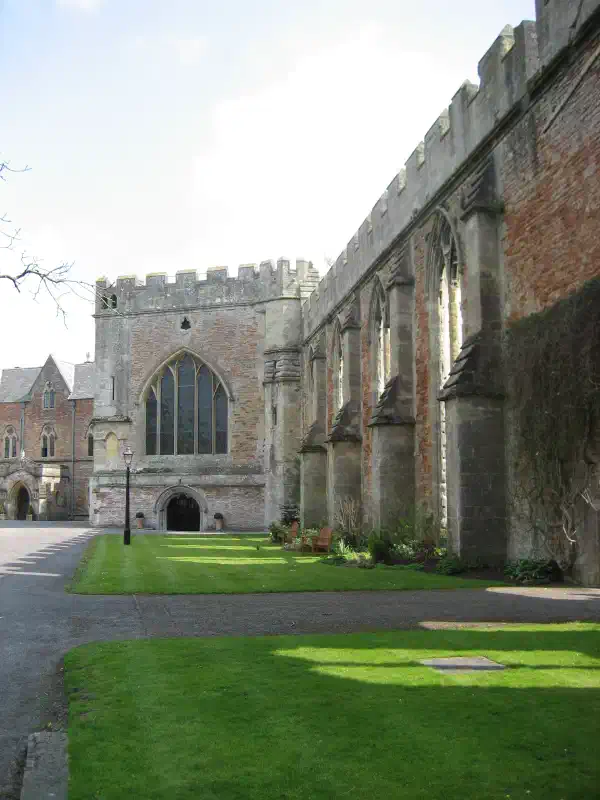 A stone wall, with a building attached in the background. In the foreground is a grassy area.